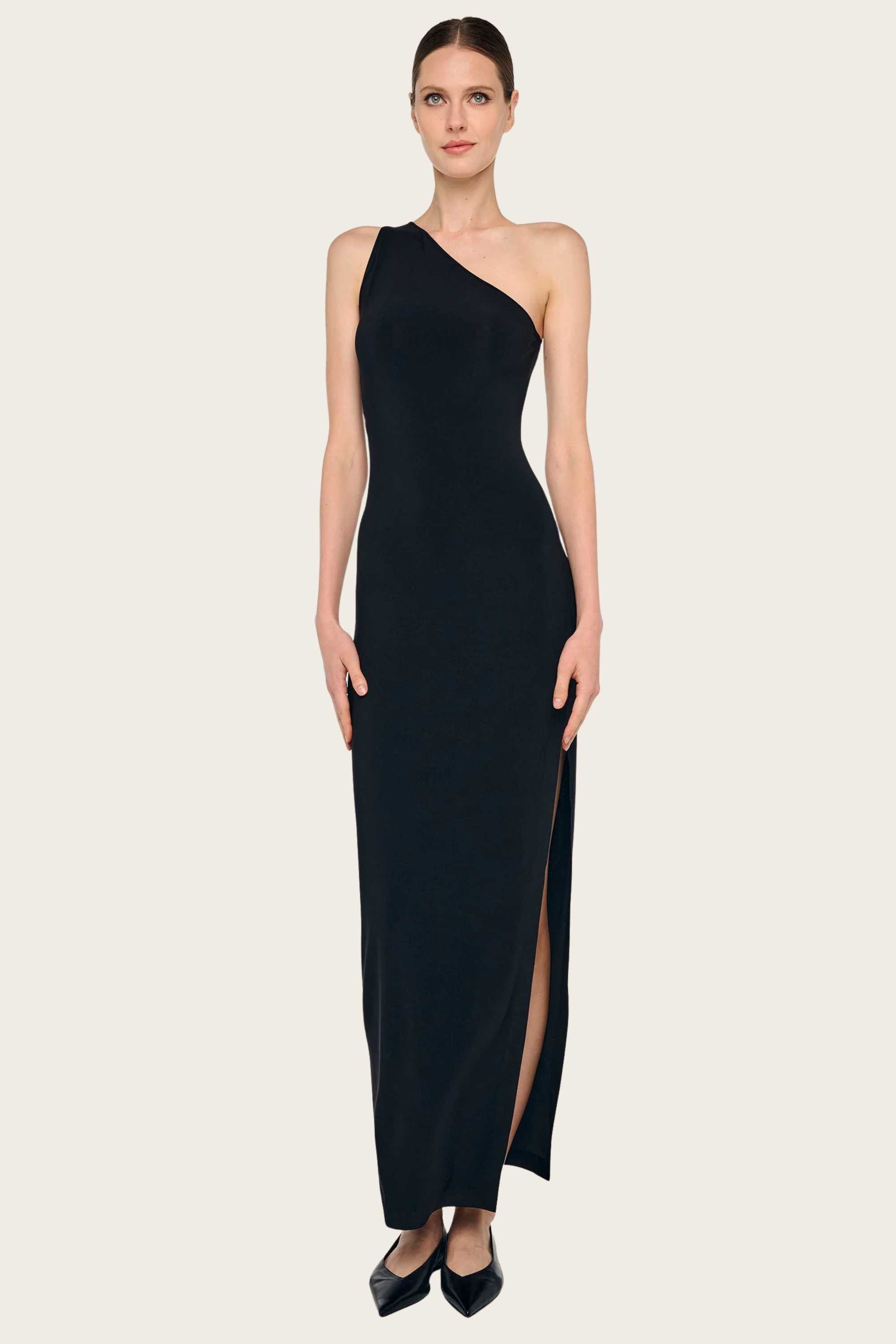 Black one-shoulder long dress with side slit by Norma Kamali, paired with black pointed-toe flats.